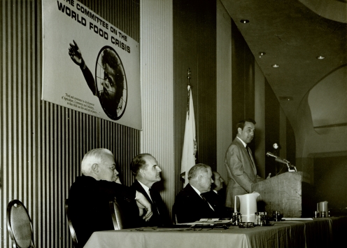 Men are seated at a table while a man speaks at a podium.