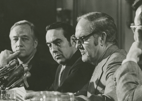 Men sit at a table with microphones pointed at one man.