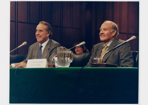 Two men smile while sitting at a table with microphones.